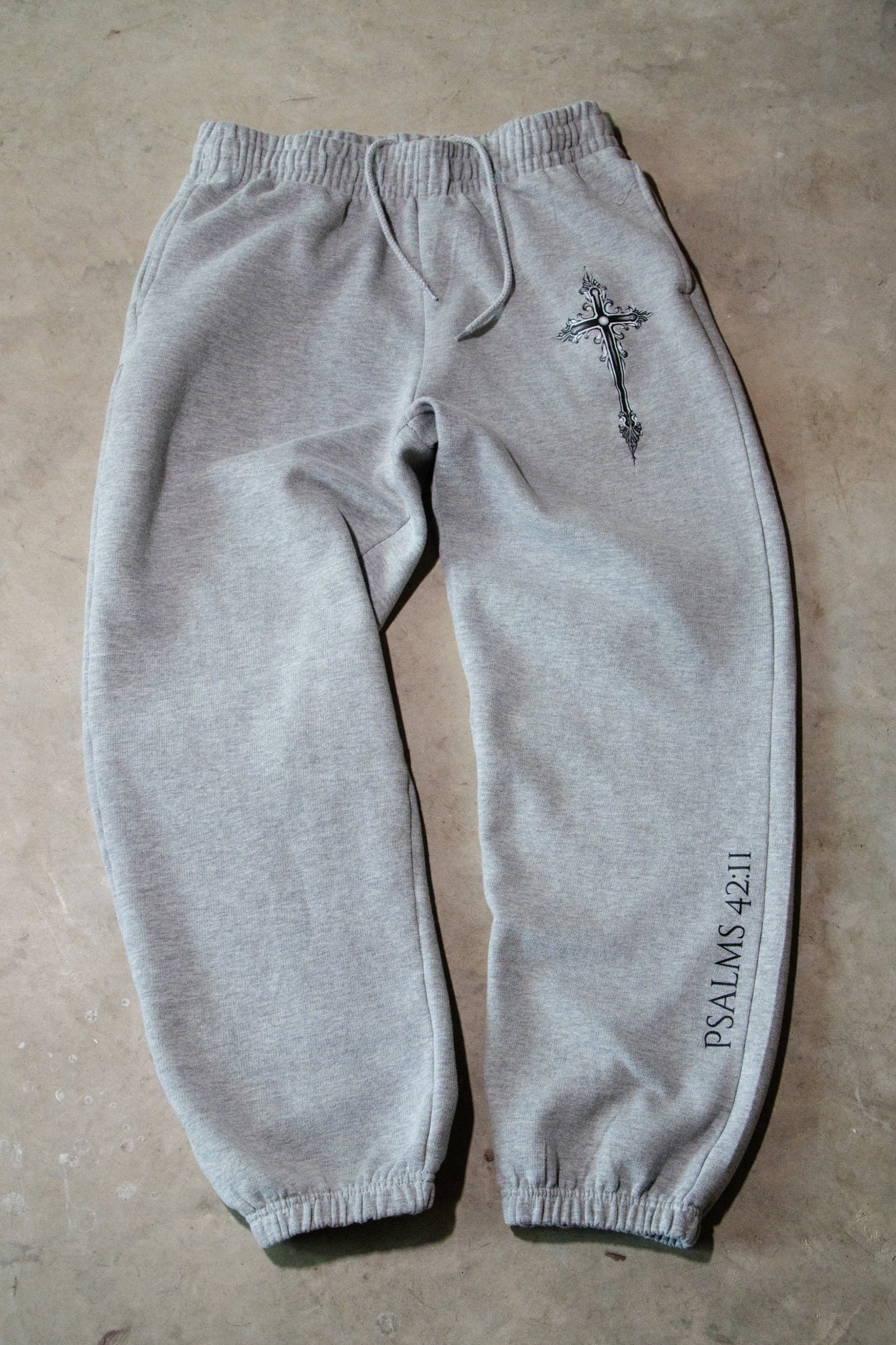 Gray sweatpants with a cross and 'Psalms 42:1' text on a beige surface